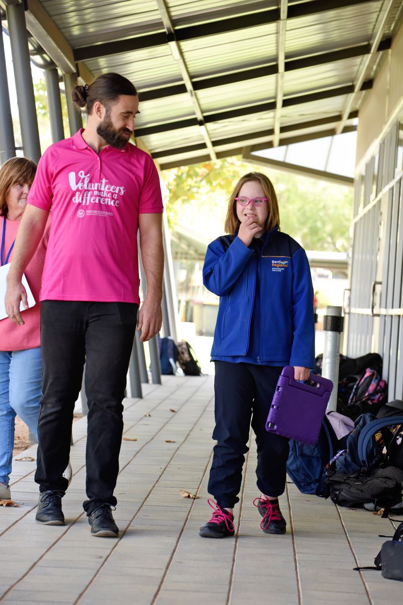 An adult volunteer walking outside with a student who is holding a tablet device.