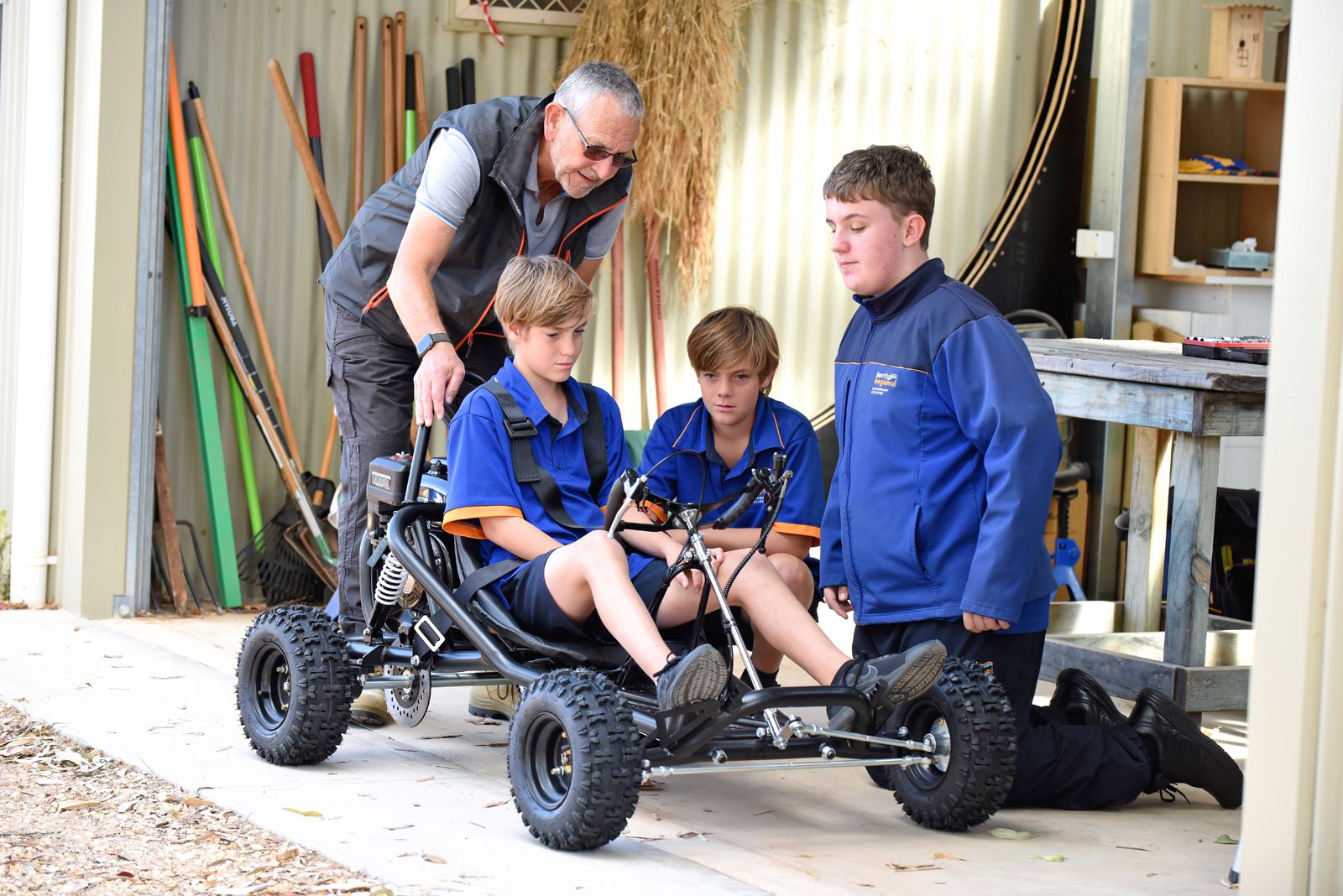 An older volunteer with 3 students, one of them is sitting in a go-kart, the other 2 are looking at it with interest.