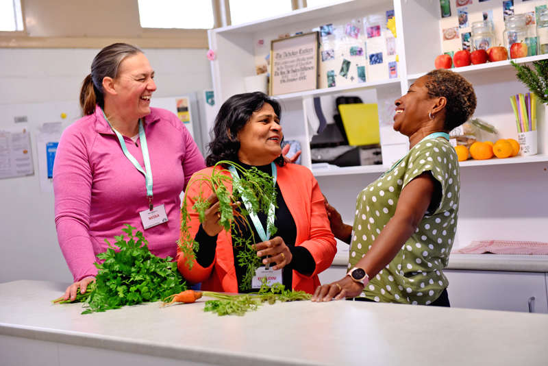 3 people at a kitchen bench, one is holding carrots with green leaves. They are laughing.
